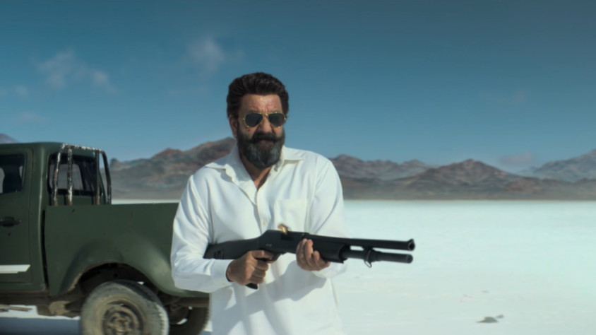 “Dhurandhar ट्रेलर का एक action सीन जिसमें सफेद कपड़ों में एक आदमी रेगिस्तान में बंदूक पकड़े खड़ा है।”
